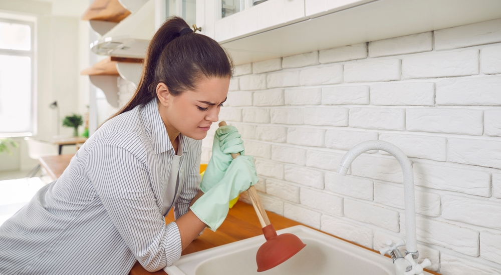 Woman having trouble using a plunger in the kitchen sink to clear a clog