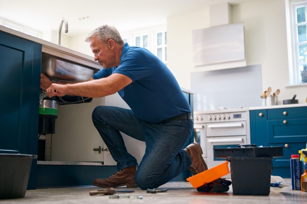 a plumber kneeling by the kitchen sink with the cabinet open as he performs an inspection and repairs on the piping